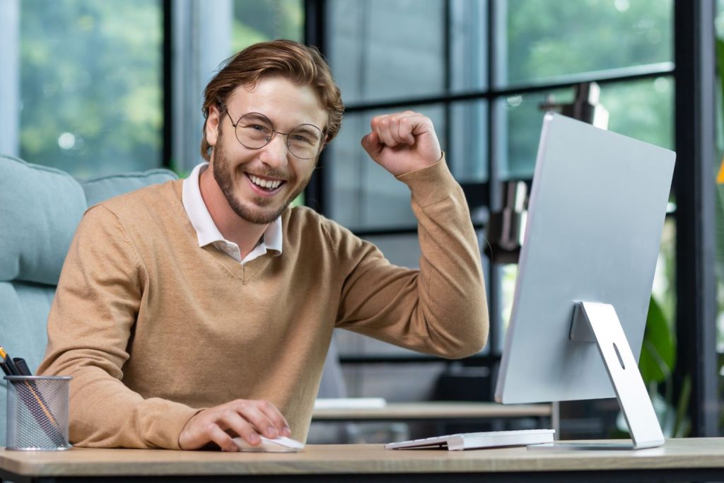 Man sitting at desk in front of a computer smiling and celebrating with hand in a fist and raised