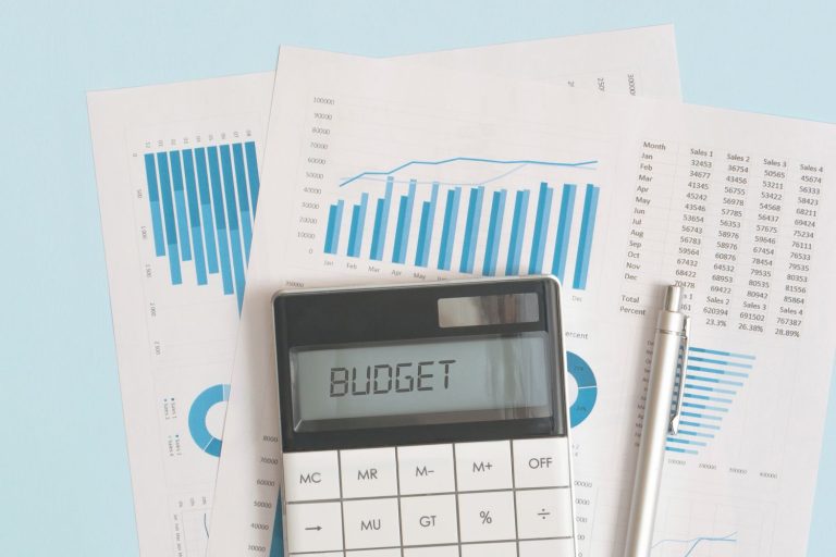 Papers with graphs and numbers with a calculator that has the word BUDGET on the screen and a pen next to it.