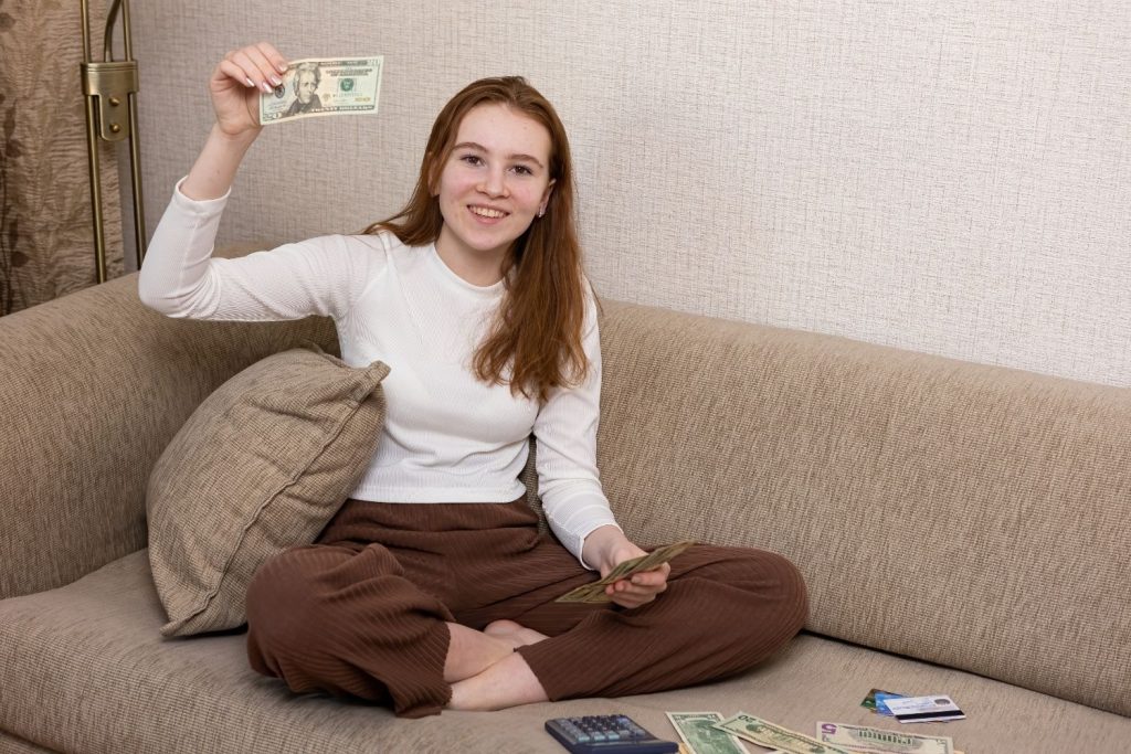 Female teen on couch with money and a calculator, holding up a dollar bill. Teaching biblical teen finance tips.