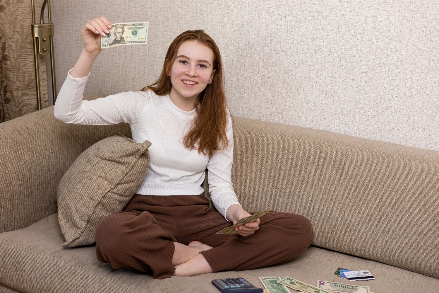 Female teen on couch with money and a calculator, holding up a dollar bill. Teaching biblical teen finance tips.