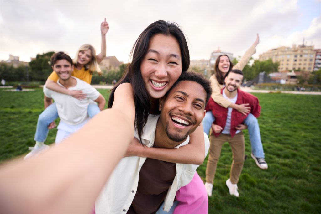 Multirracial group of friends taking selfie picture in a park with couples hugging and celebrating with arms up. One Another Bible Verses